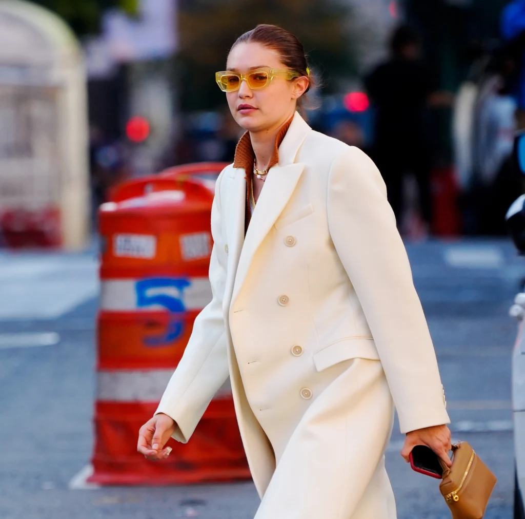 Kobieta w długim kremowym płaszczu i żółtych okularach przeciwsłonecznych spaceruje po miejskiej ulicy, trzymając w ręku małą brązową torebkę; w tle widoczne są biało-pomarańczowe przeszkody drogowe.