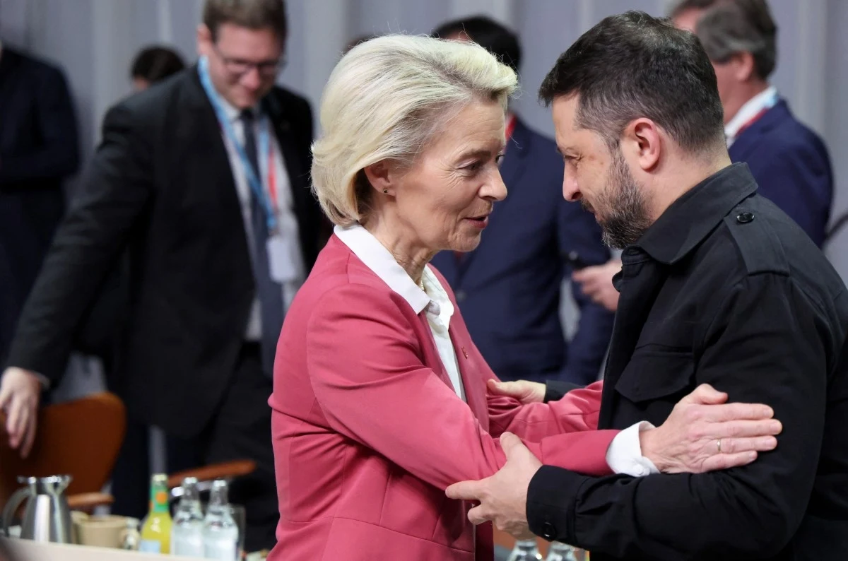 Przewodnicząca Komisji Europejskiej Ursula von der Leyen i prezydent Ukrainy Wołodymyr Zełenski Dwie osoby w serdecznym uścisku wymieniające uprzejmości podczas oficjalnego spotkania, w tle kilku innych uczestników wydarzenia przy stole konferencyjnym.