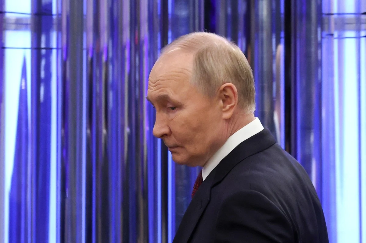 Prezydent Rosji Władimir Putin Mężczyzna w garniturze o jasnych włosach ukazany z profilu, na tle połyskujących, niebiesko-fioletowych pionowych paneli.