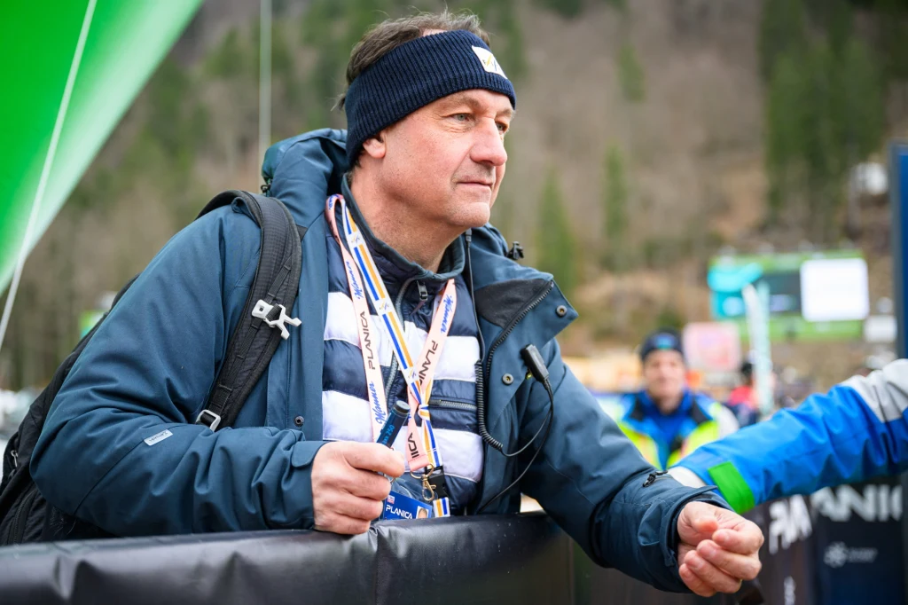 Mężczyzna w niebieskiej kurtce i opasce na głowie stoi przy ogrodzeniu z identyfikatorami na szyi, z tyłu rozmyty krajobraz górski oraz osoby w kurtkach sportowych.