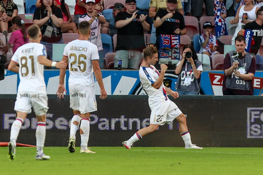 Piłkarze Górnika Zabrze w meczu Ekstraklasy Piłkarz drużyny w białych strojach wykonuje charakterystyczny gest po zdobyciu gola, otoczony przez kolegów z zespołu oraz dopingowaną publiczność na trybunach; w tle widać kibiców i fotoreporterów.