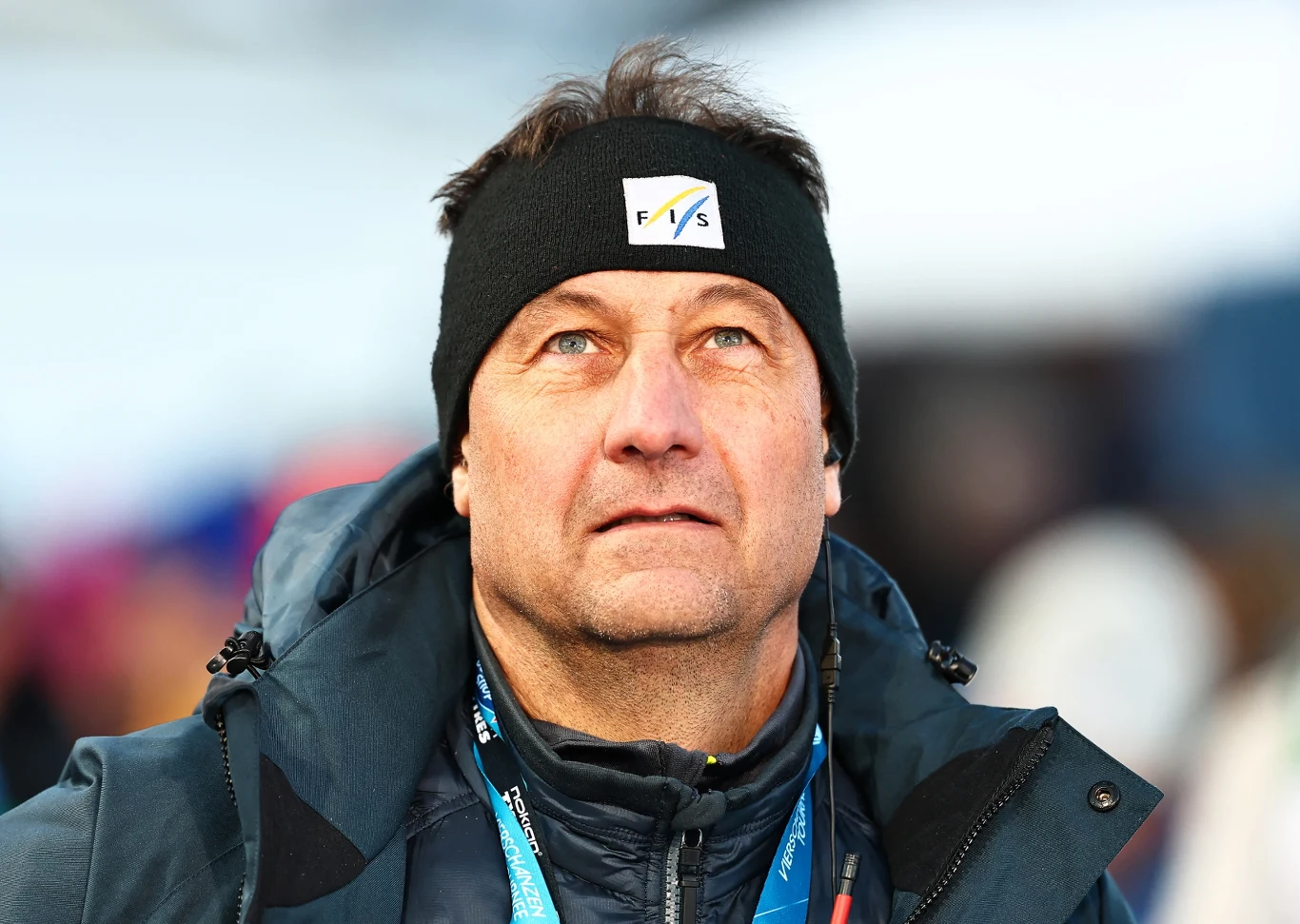 Sandro Pertile, dyrektor Pucharu Świata Mężczyzna w średnim wieku ubrany w sportową kurtkę i czapkę z logo FIS, patrzy lekko w górę, tło rozmyte z kolorowymi plamami sugerującymi obecność innych ludzi.