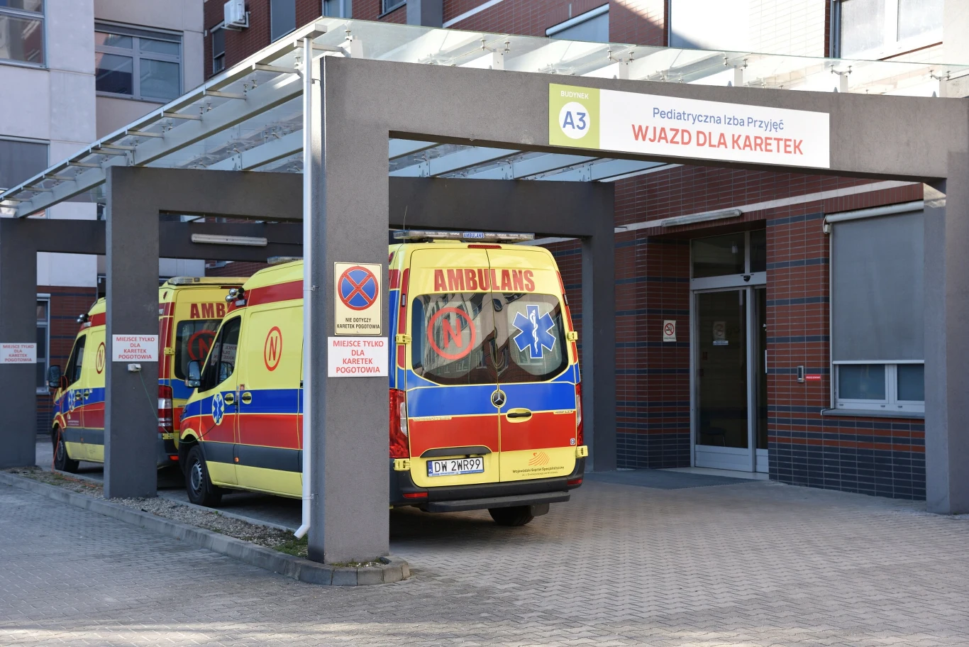 Apel do premiera w sprawie zabezpieczenia pieniędzy na leczenie pacjentów w publicznym systemie Dwa ambulansy zaparkowane pod zadaszeniem przy wejściu oznaczonym jako pediatryczna izba przyjęć, wjazd dla karetek, przy budynku szpitala, tablica informacyjna oraz zakaz postoju.