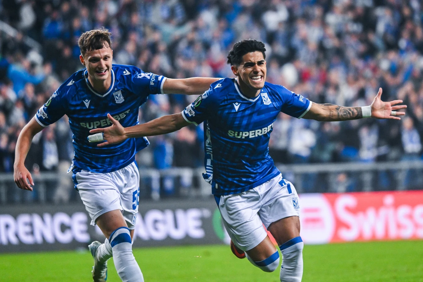 Dwóch piłkarzy w niebieskich strojach świętuje zdobytego gola na boisku, kibice w tle tworzą sportową atmosferę.