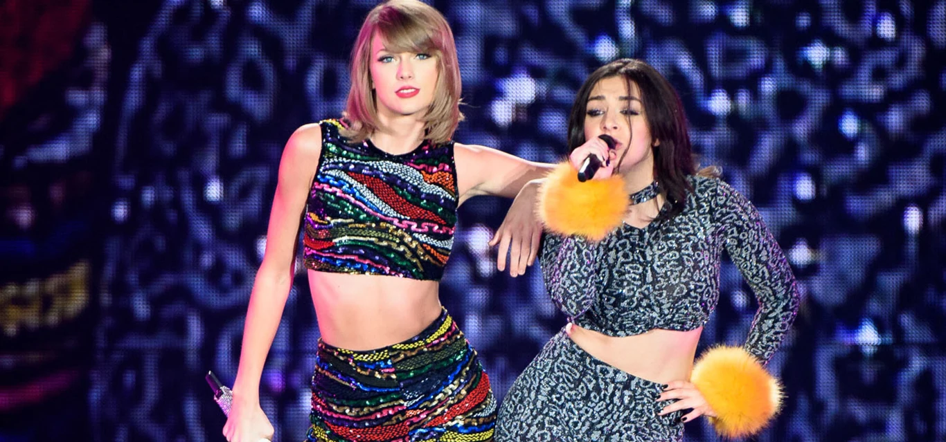 Taylor Swift wydała album "The Life of a Showgirl" Dwie młode kobiety śpiewające razem na scenie, jedna w kolorowym, błyszczącym zestawie top i spódniczka, druga w szarym stroju z wzorem i żółtymi puszystymi mankietami, w tle dynamiczne, kolorowe światła i wzory.