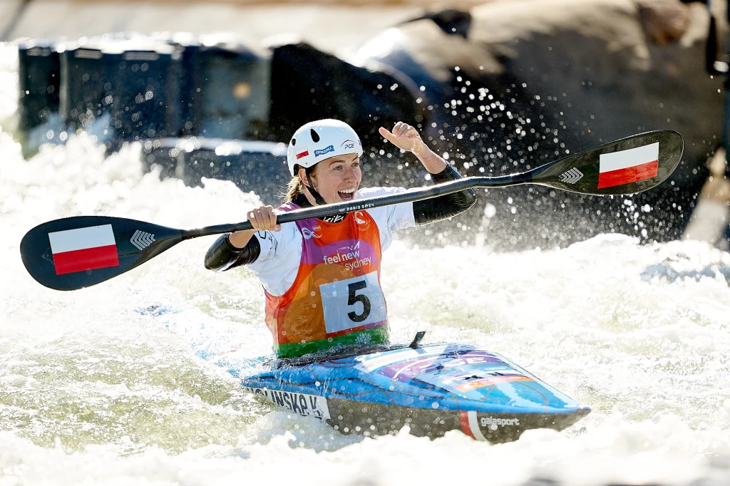 Klaudia Zwolińska Kajakarka w kamizelce z numerem startowym pokonuje wodny tor slalomowy, unosząc w geście triumfu wiosło z flagą Polski, twarz wyraża radość i zadowolenie z osiągniętego wyniku
