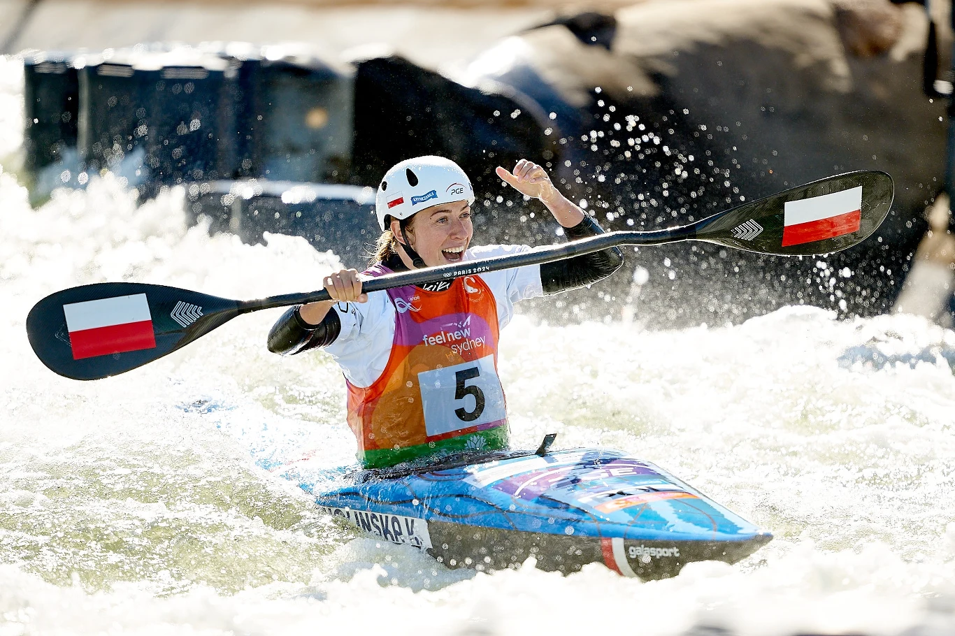 Kajakarka w kamizelce z numerem startowym pokonuje wodny tor slalomowy, unosząc w geście triumfu wiosło z flagą Polski, twarz wyraża radość i zadowolenie z osiągniętego wyniku