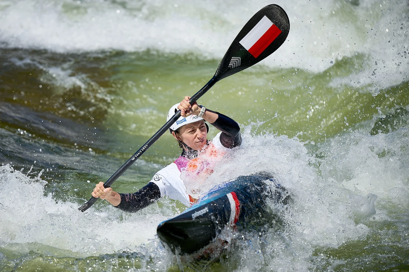 Kajakarz w trakcie dynamicznego spływu przez gwałtowne fale na rzece, skupiony na sterowaniu, kajak oznaczony polskimi barwami narodowymi, intensywna akcja i bryzgi wody wokół.