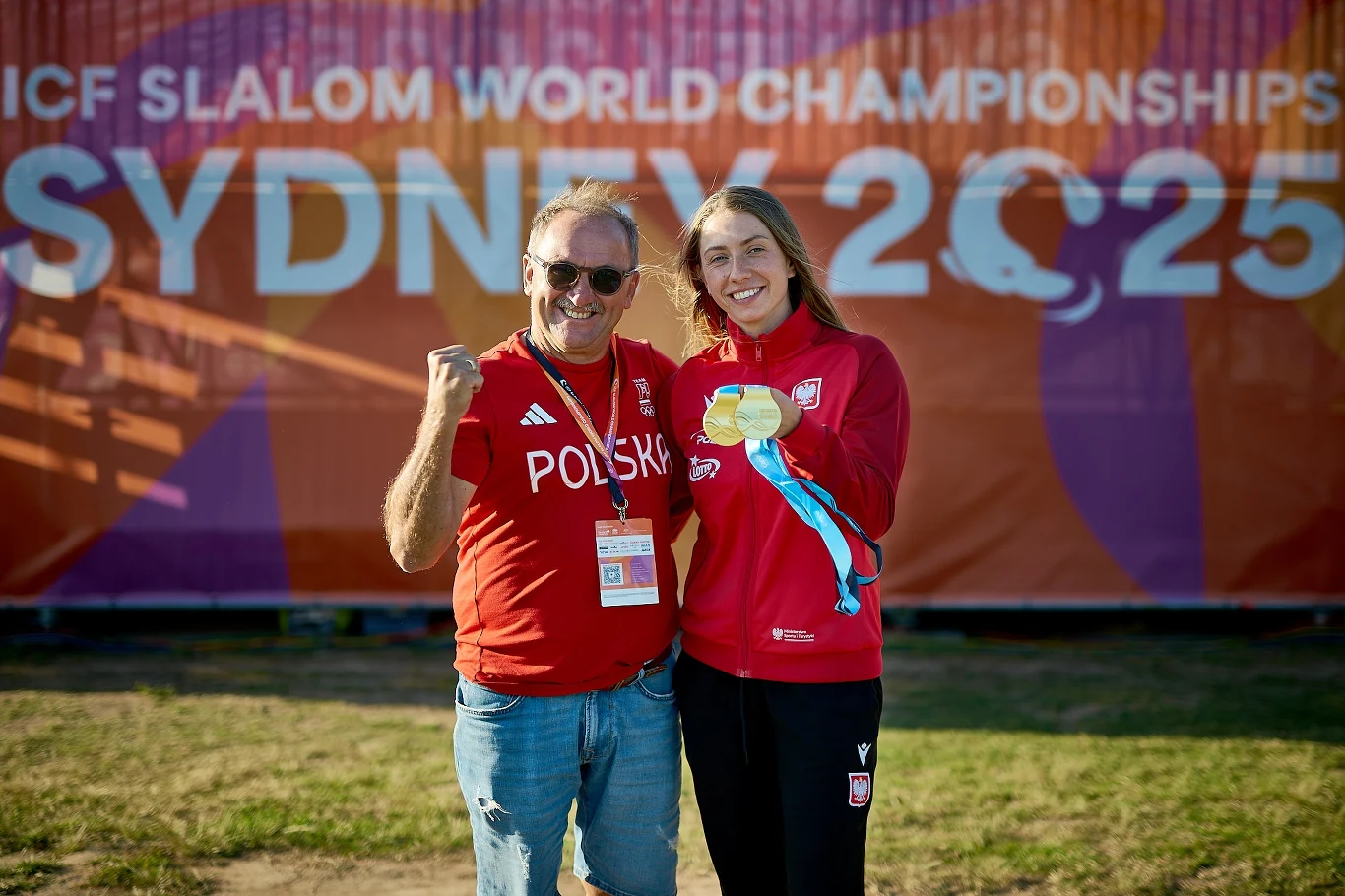 Dwie uśmiechnięte osoby w czerwonych strojach z napisem 'Polska', stoją na tle banera z napisem 'ICF Slalom World Championships Sydney 2025', jedna z osób prezentuje złoty medal.