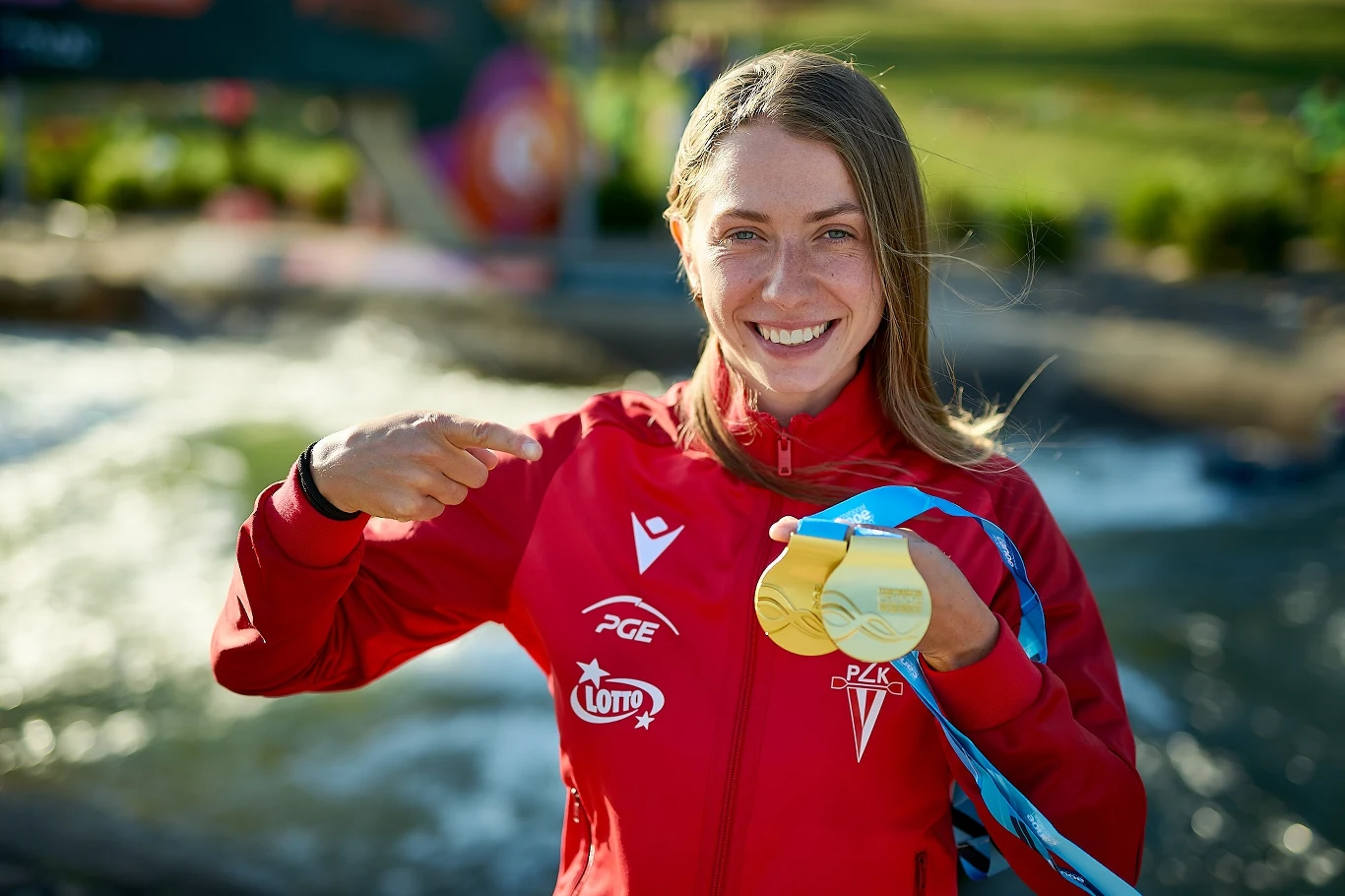 Młoda sportsmenka w czerwonym dresie z logo sponsorów uśmiecha się i dumnie wskazuje na złoty medal zawieszony na szyi, w tle rozmyte otoczenie na świeżym powietrzu.