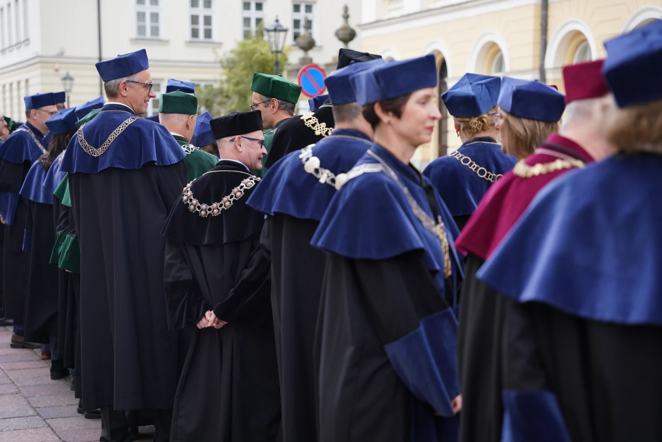 Czy studiowanie się opłaca? Komisja Europejska wskazuje różnice w zarobkach. Grupa osób w tradycyjnych togach i biretach akademickich stojąca w szeregu na zewnątrz budynku, ozdobiona łańcuchami rektorskimi.