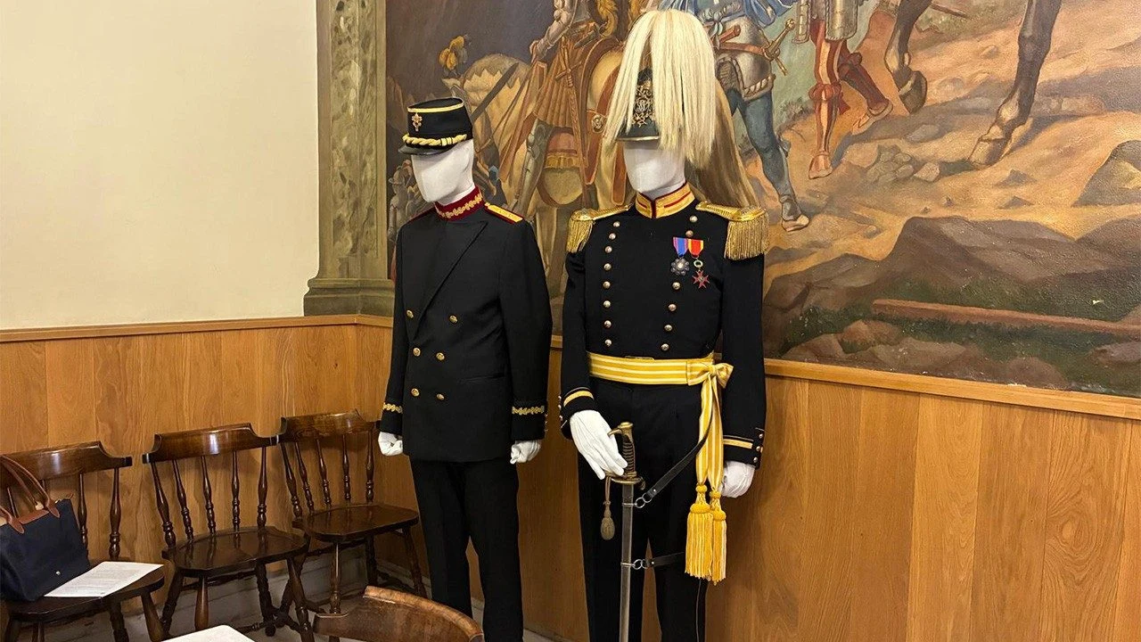 Dwa manekiny ubrane w historyczne mundury wojskowe stoją na tle ściany pokrytej malowidłem przedstawiającym scenę z udziałem jeźdźców na koniach; obok ustawione są drewniane krzesła.