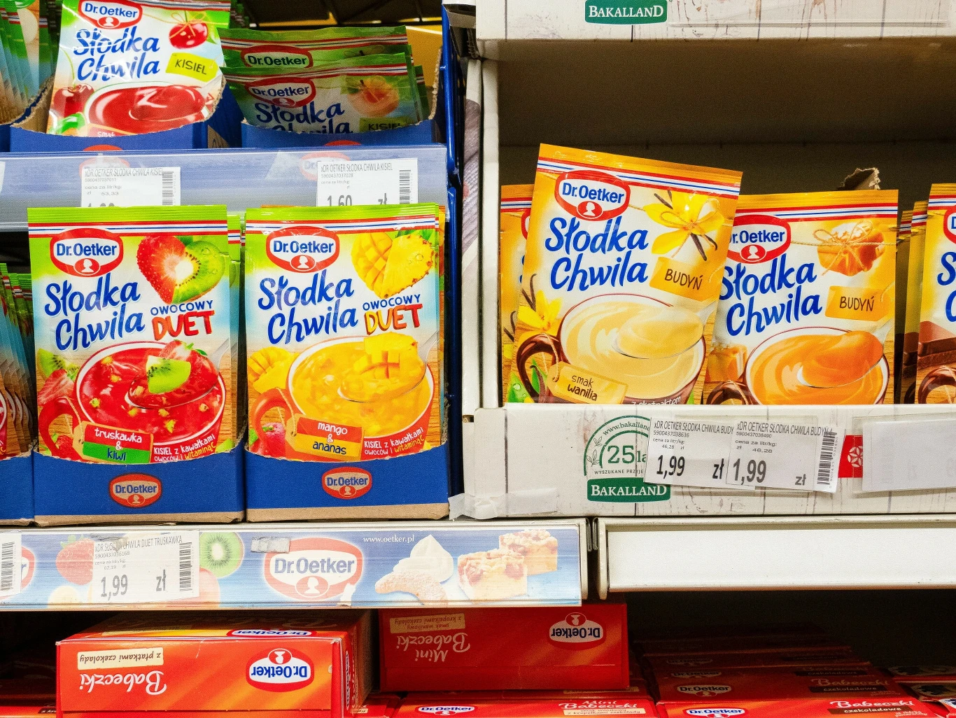 Opakowania po produktach sypkich zazwyczaj wrzucamy do żółtego pojemnika Opakowania produktów instant do przygotowania deserów marki Dr. Oetker o smakach owocowych i budyniowych ułożone na półce sklepowej, widoczne ceny i etykiety.