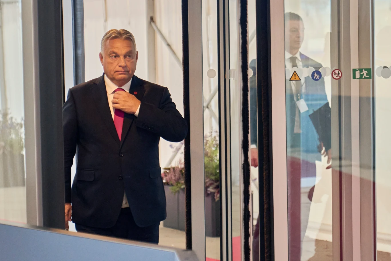 Premier Węgier Viktor Obran Mężczyzna w garniturze z czerwonym krawatem przechodzi przez drzwi obrotowe w nowoczesnym wnętrzu. W tle widoczny inny mężczyzna ubrany w garnitur oraz elementy architektury i oznaczenia informacyjne na szkle.
