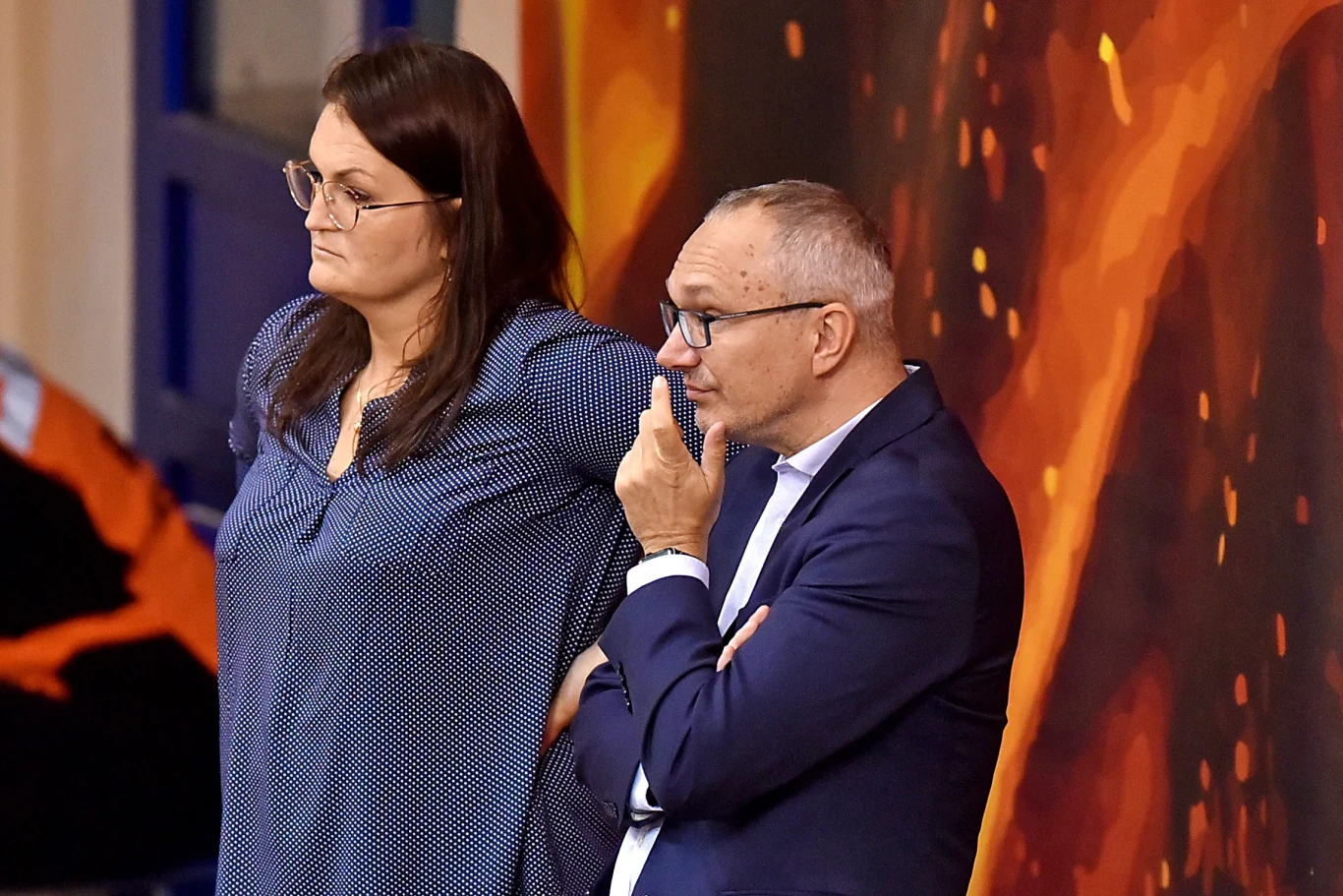Dwoje dorosłych, elegancko ubranych ludzi stojących obok siebie, kobieta w granatowej bluzce i mężczyzna w garniturze, oboje patrzą w jednym kierunku z poważnymi minami, w tle barwna, abstrakcyjna ściana.