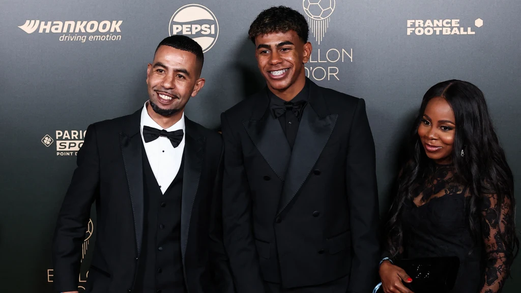 Lamine Yamal z rodzicami Trzy elegancko ubrane osoby pozują na tle ściany z logotypami wydarzenia, uśmiechając się i patrząc w stronę aparatu.