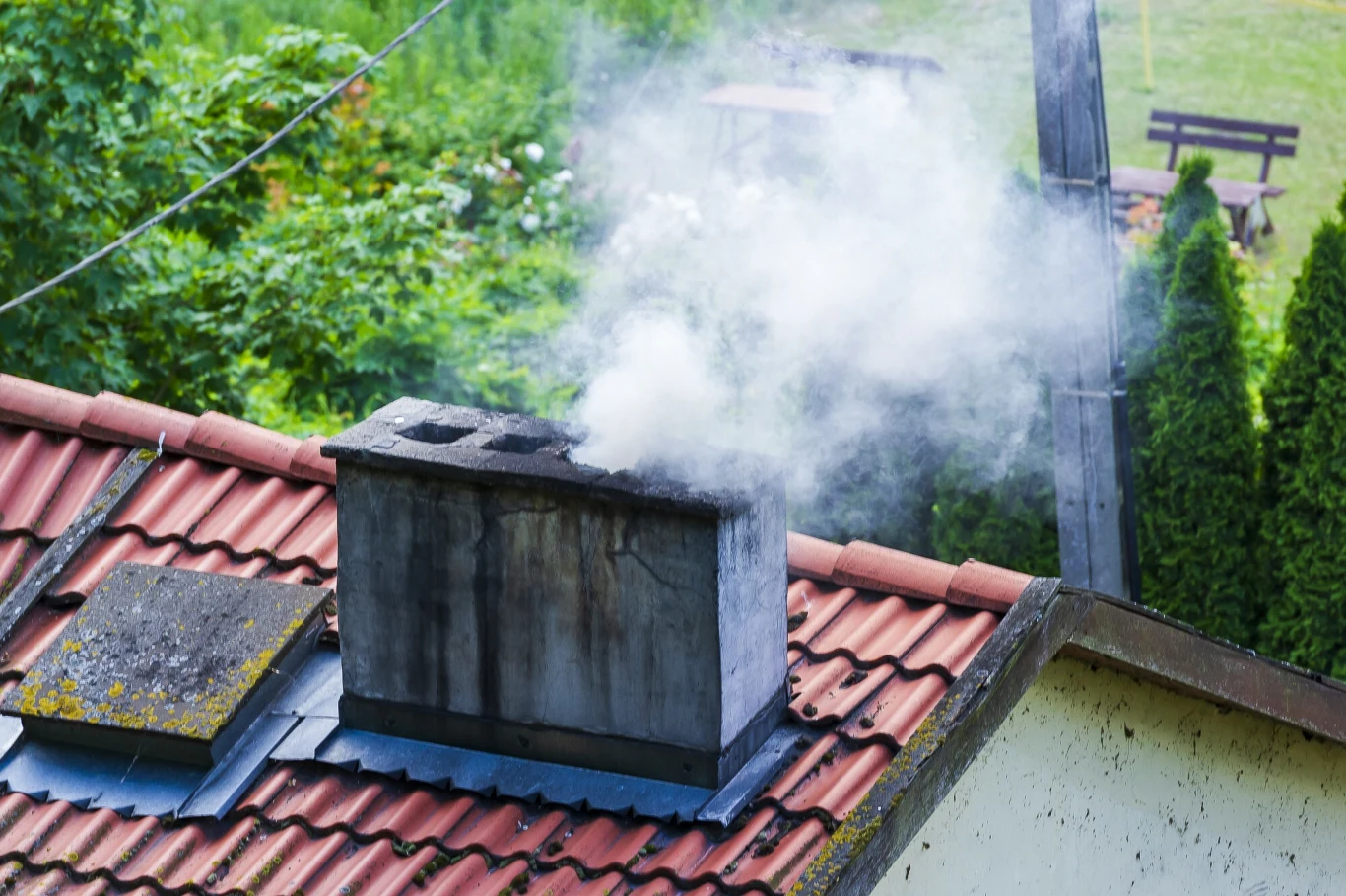 murowany komin na skośnym dachu pokrytym dachówką, z którego wydobywa się biały dym, w tle zielone drzewa i ogród