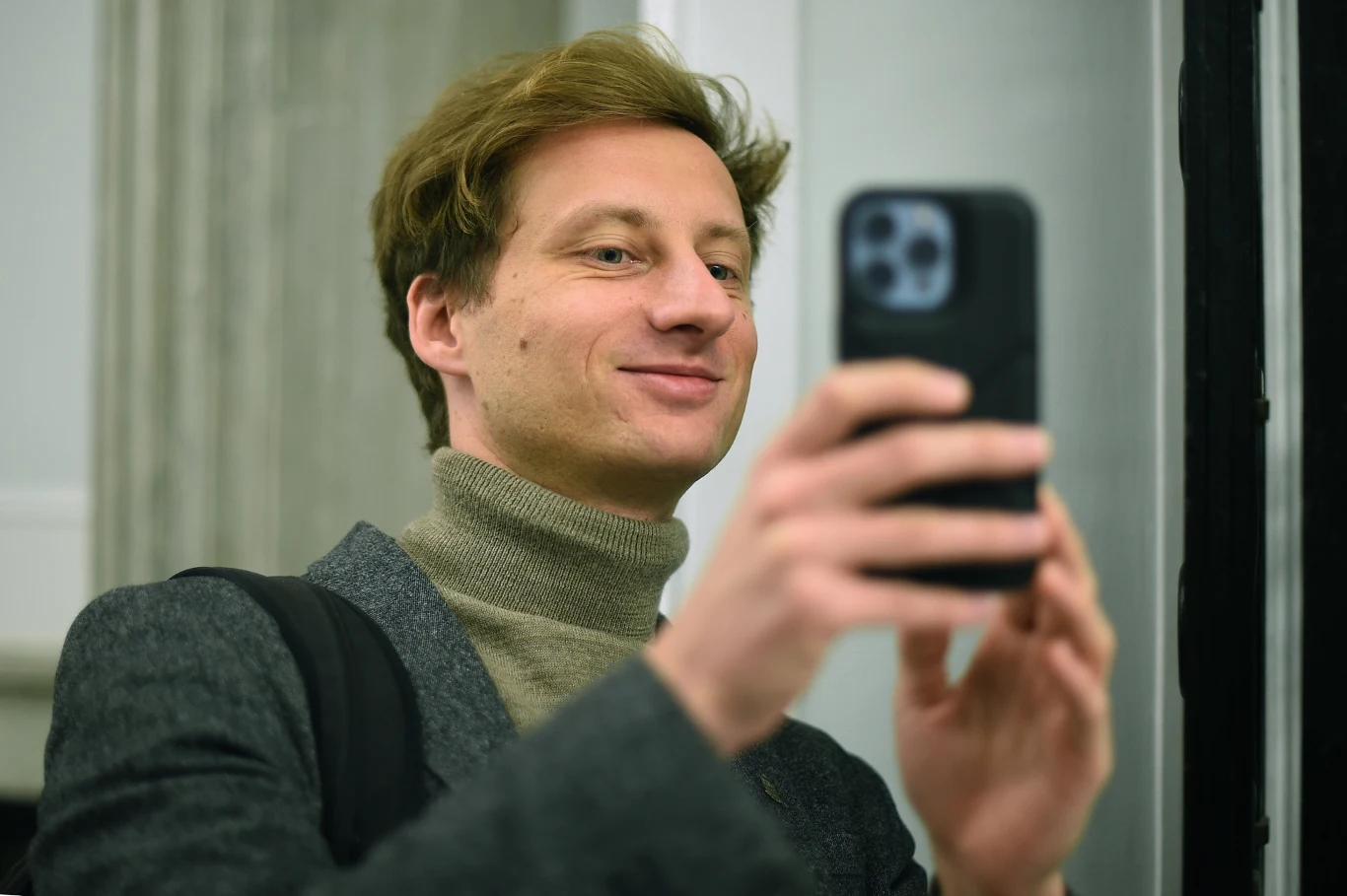 Franciszek Sterczewski Mężczyzna w szarym swetrze oraz marynarce robi selfie smartfonem, lekko się uśmiecha, tło stanowią jasne, neutralne ściany.