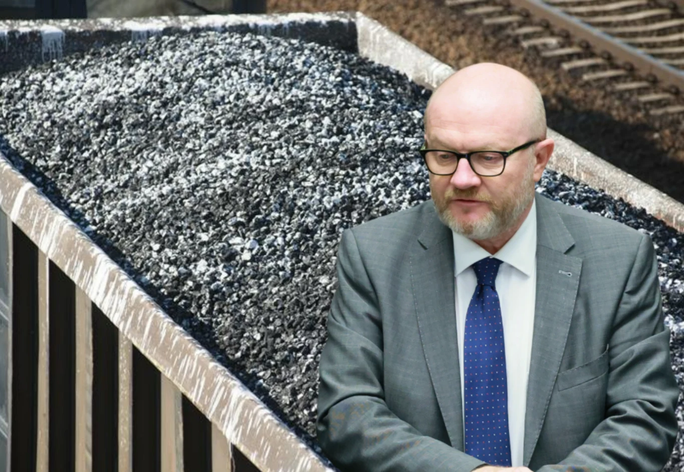 Wiceszef MSWiA Maciej Duszczyk komentuje ostatni incydent z odczepieniem wagonu w Katowicach Mężczyzna w garniturze i okularach stoi przed węglarką wypełnioną węglem, w tle widoczne są tory kolejowe.