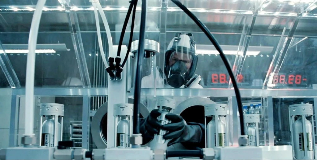 Pracownik laboratorium ubrany w odzież ochronną i maskę pracuje przy zaawansowanej aparaturze laboratoryjnej w sterylnym otoczeniu, otoczony przez przezroczyste osłony i widoczne liczniki LED.
