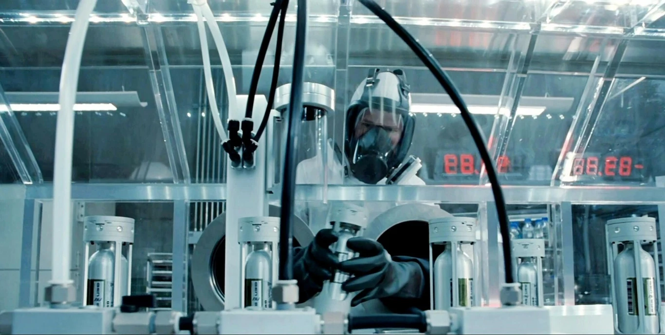 Pracownik laboratorium ubrany w odzież ochronną i maskę pracuje przy zaawansowanej aparaturze laboratoryjnej w sterylnym otoczeniu, otoczony przez przezroczyste osłony i widoczne liczniki LED.