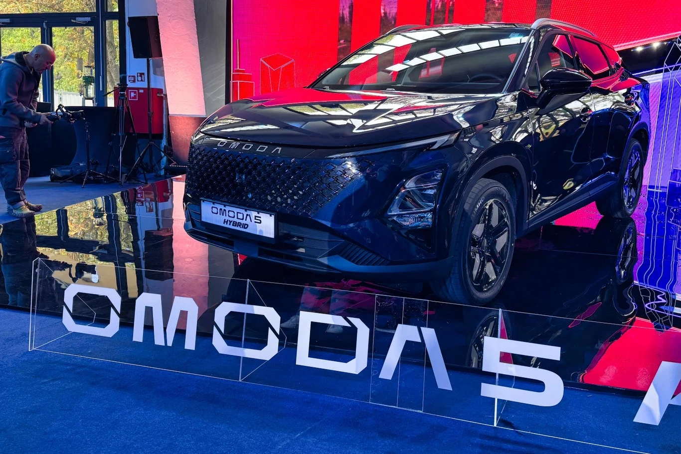 Do wersji benzynowej i elektrycznej dołącza Omoda 5 Hybrid Elektryczny samochód Omoda 5 Hybrid prezentowany na ekspozycji targowej, ustawiony na podwyższeniu z efektownym oświetleniem i wyeksponowanym logo marki. W tle elementy scenografii targowej i osoby zwiedzające.