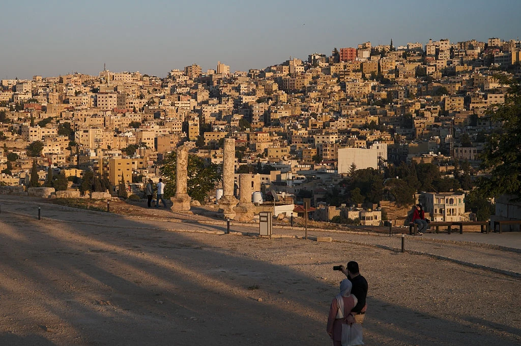 Amman - stolica Jordanii (zdj. ilustracyjne) Panorama miasta z widocznymi licznymi budynkami, na pierwszym planie starożytne ruiny z trzema kamiennymi kolumnami oraz niewielka liczba ludzi spacerujących w pobliżu.