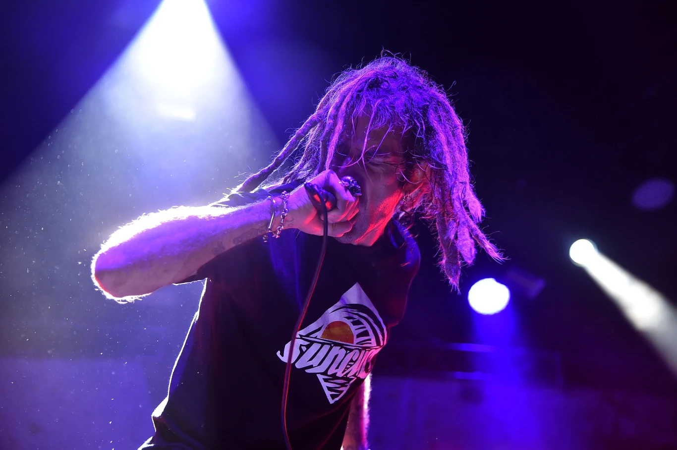 Randy Blythe, wokalista Lamb of God Mężczyzna z dredami śpiewający energicznie do mikrofonu na scenie koncertowej, otoczony kolorowymi światłami scenicznymi, w koszulce z wyrazistym nadrukiem.