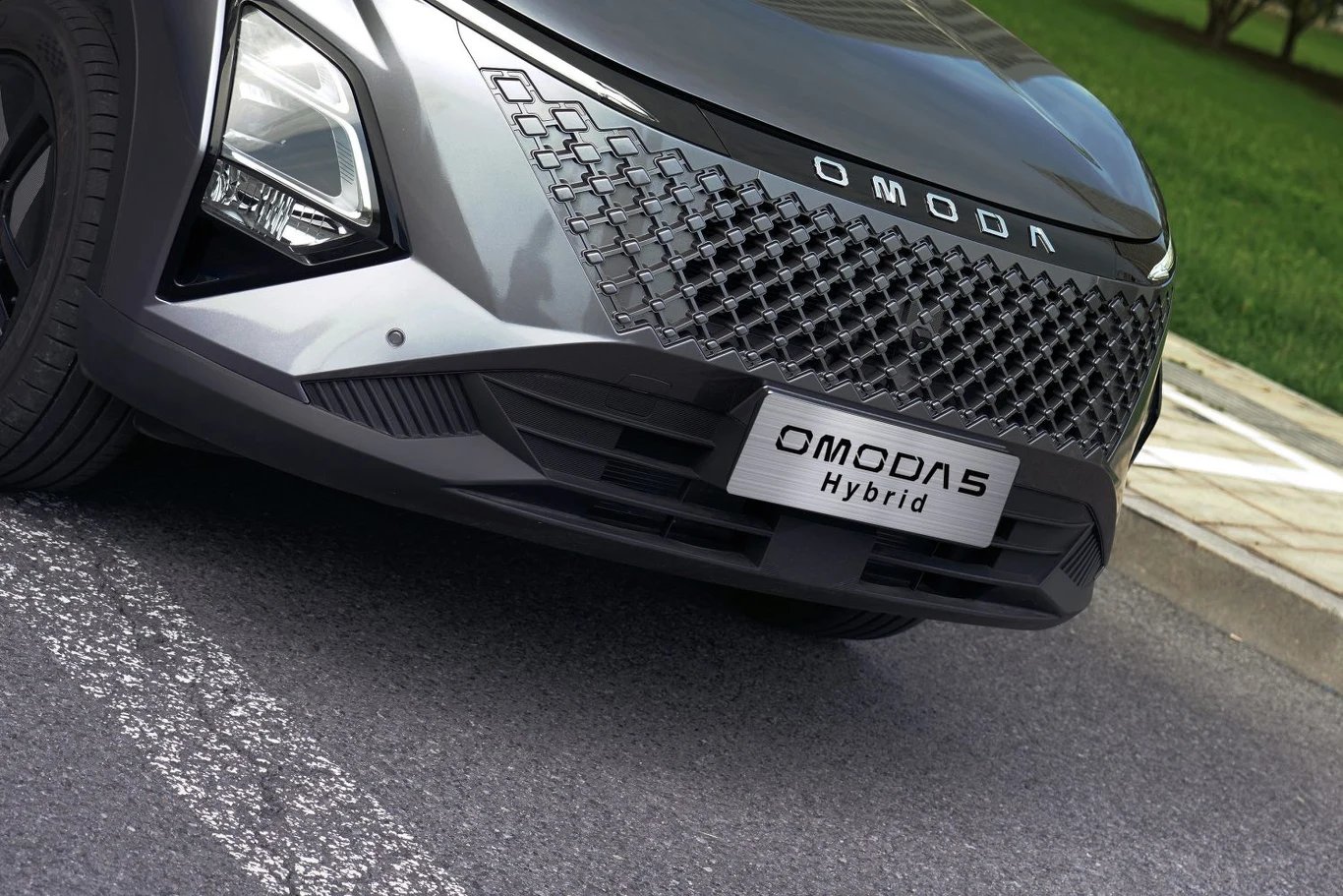 Auto przeszło drobny lifting - odświeżono grill i reflektory Nowoczesny przód samochodu Omoda Hybrid z charakterystycznym grillem i wyraźnie widoczną tablicą rejestracyjną, częściowo zaparkowanego na parkingu przy trawie.