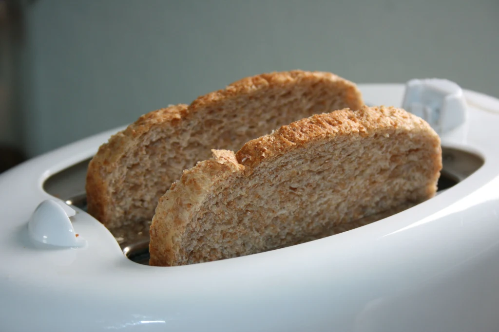 Toster też może okazać się warty więcej niż myślisz Dwie kromki chleba pełnoziarnistego umieszczone w białym tosterze, gotowe do opiekania, widoczne z bliska pokrętła urządzenia.