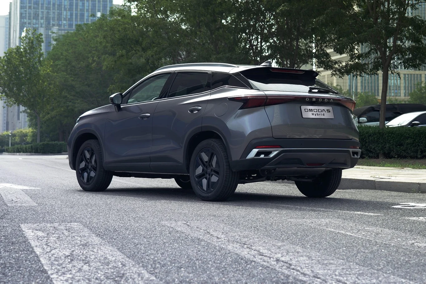 Z tyłu hybrydowa Omoda nie różni się od spalinowej odpowiedniczki Nowoczesny SUV w kolorze grafitowym stoi na pustej ulicy miejskiej, w otoczeniu zieleni, z widoczną tablicą rejestracyjną i nowoczesnym designem nadwozia.