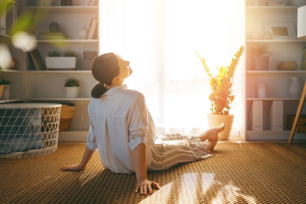 Ten trik jest tani, ale w Polsce mało znany. Dzięki niemu w twoim domu będzie wyjątkowo pachnieć Kobieta siedząca na podłodze w jasnym, przytulnym pokoju, oparta o ręce i patrząca w kierunku wpadającego przez duże okno światła słonecznego, w otoczeniu roślin doniczkowych oraz półek z książkami.