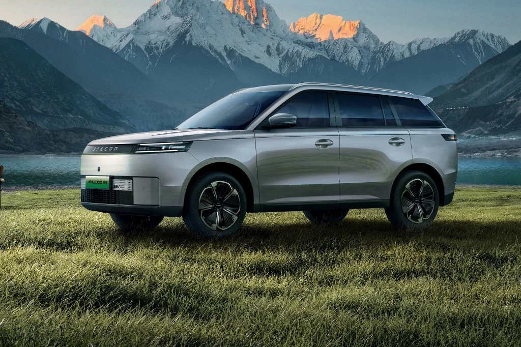 Srebrny nowoczesny SUV stoi na zielonej trawie z malowniczym jeziorem oraz ośnieżonymi górskimi szczytami w tle, jasne światło poranka podkreśla kształty pojazdu.