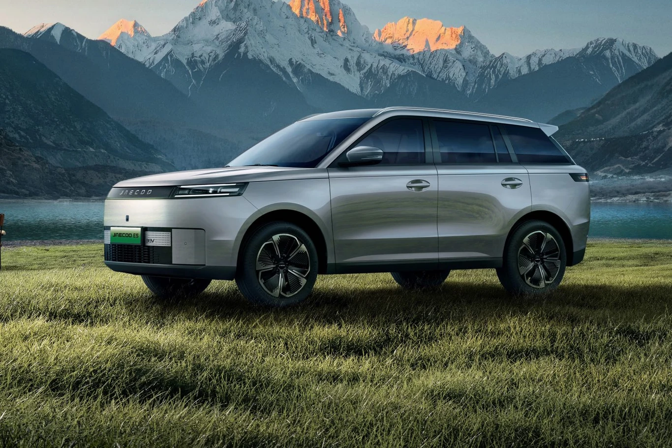 Srebrny nowoczesny SUV stoi na zielonej trawie z malowniczym jeziorem oraz ośnieżonymi górskimi szczytami w tle, jasne światło poranka podkreśla kształty pojazdu.