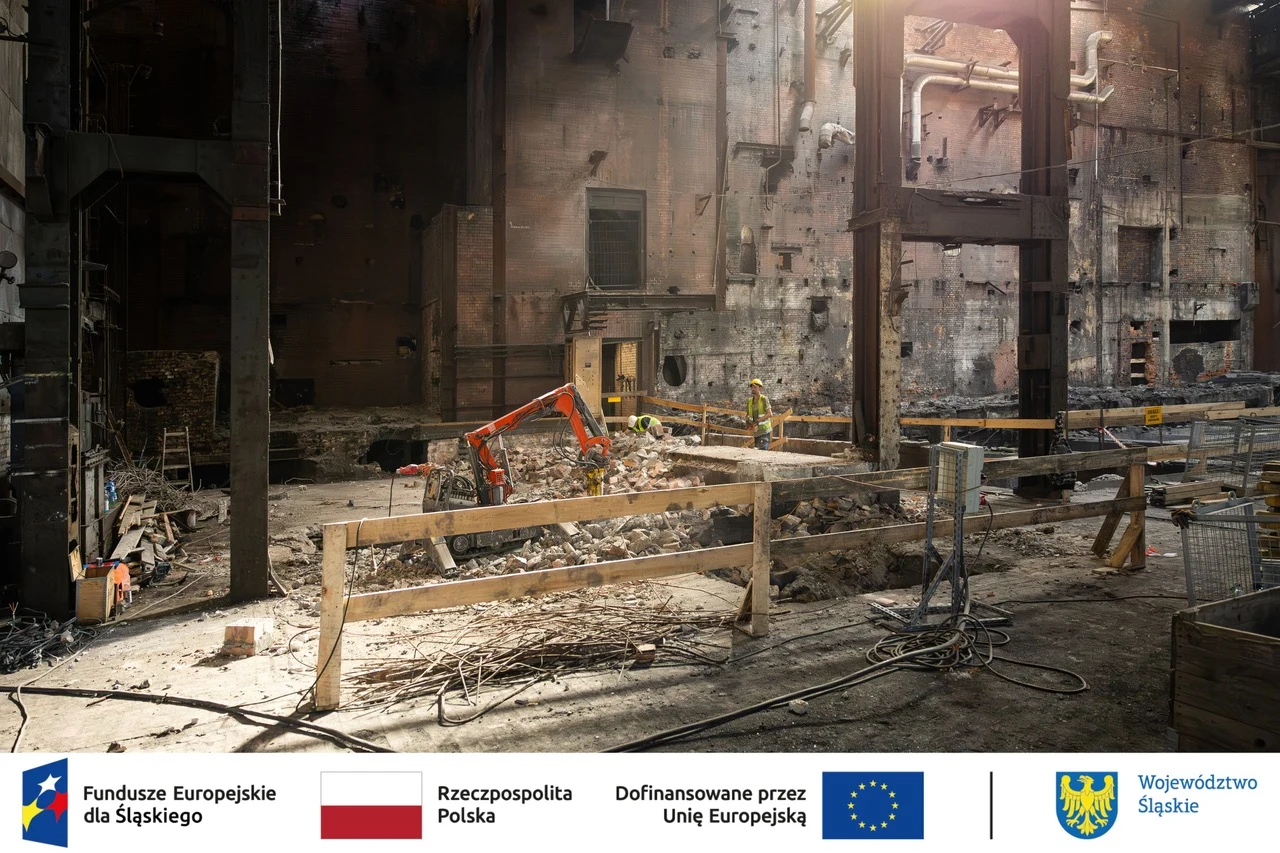 Opuszczona hala przemysłowa z widocznymi elementami rozbiórki, resztkami gruzu i barierkami zabezpieczającymi teren prac. Na środku znajdują się dwa niewielkie sprzęty budowlane, a całość oświetlona jest naturalnym światłem wpadającym przez górne otwory.