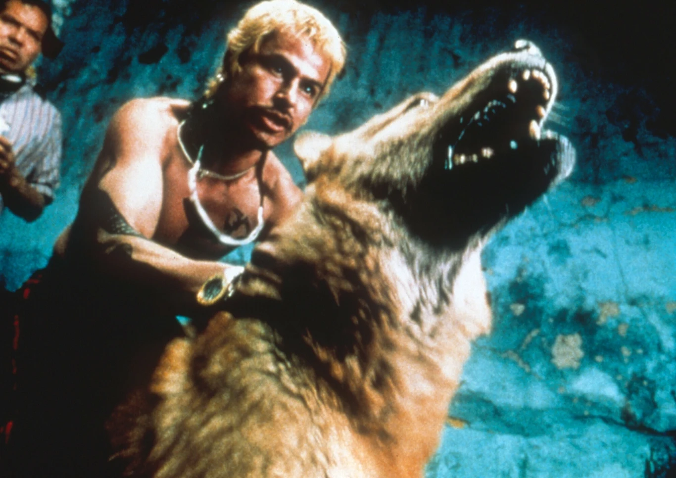 Kadr z "Amores perros" Mężczyzna o jasnych włosach i wyrazistym spojrzeniu trzyma dużego, groźnie wyglądającego wilka z wyszczerzonymi zębami. Obaj znajdują się na tle niebieskiej ściany, co podkreśla intensywność sceny.
