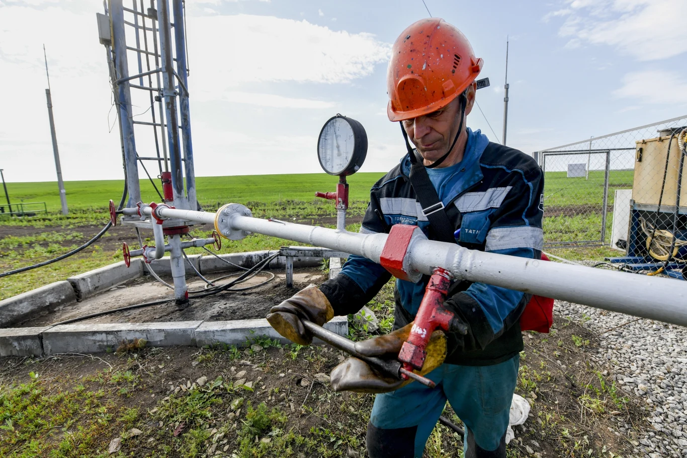 Rosyjski sektor produkcyjny zaliczył czwarty miesiąc z rzędu ze spadkiem aktywności biznesowej Mężczyzna w kasku ochronnym i roboczym stroju obsługuje metalową instalację przemysłową na tle pola i infrastruktury technicznej.