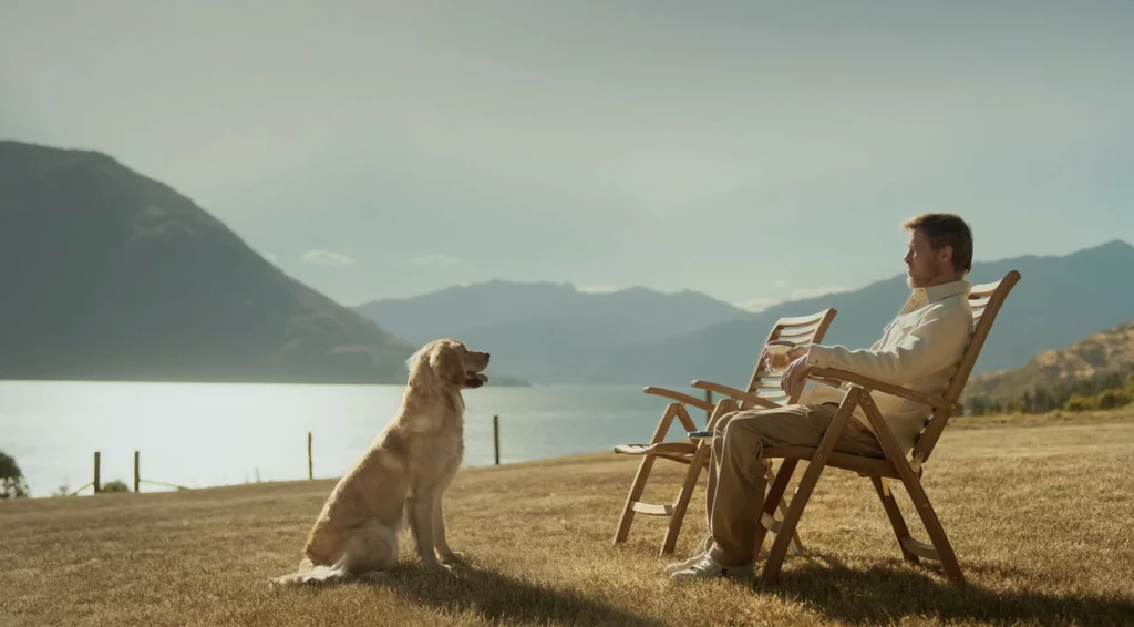 Mężczyzna w jasnych ubraniach siedzi na drewnianym krześle na suchej trawie nad jeziorem, obok niego na trawie siedzi pies rasy golden retriever, w tle rozległa panorama gór i spokojne jezioro.