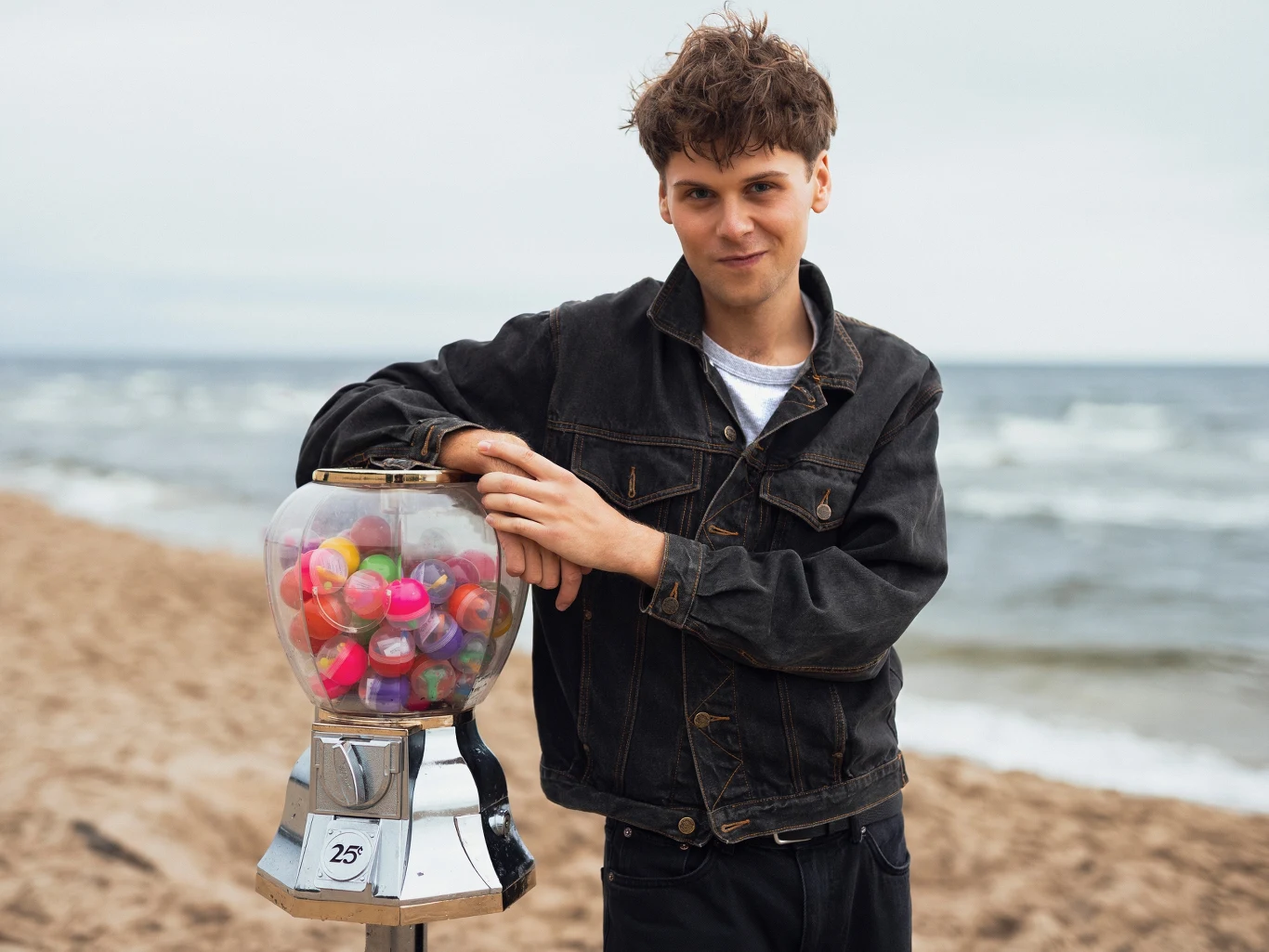 Jan Marczewski przygotował nowy materiał Młody mężczyzna w czarnej kurtce jeansowej opiera się o automat na kapsułki z zabawkami umieszczony na piaszczystej plaży nad morzem, w tle fale i pochmurne niebo.