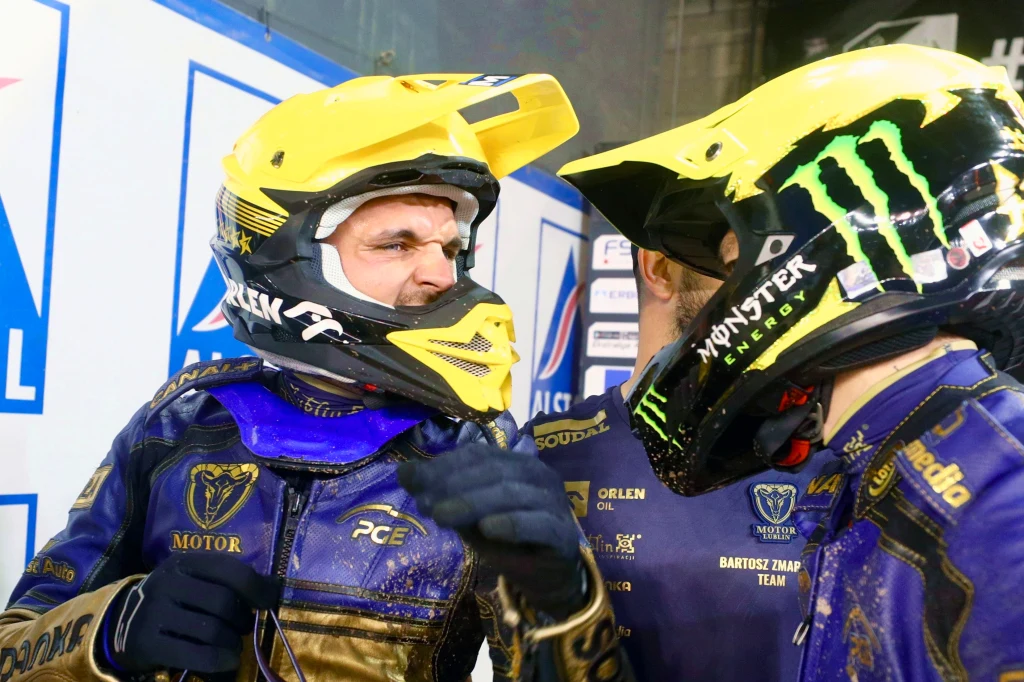 Orlen Oil Motor Lublin Dwóch motocyklistów ubranych w stroje wyścigowe z żółtymi kaskami rozmawia ze sobą, obaj mają na sobie kombinezony w barwach zespołu oraz widoczne logo sponsorów.