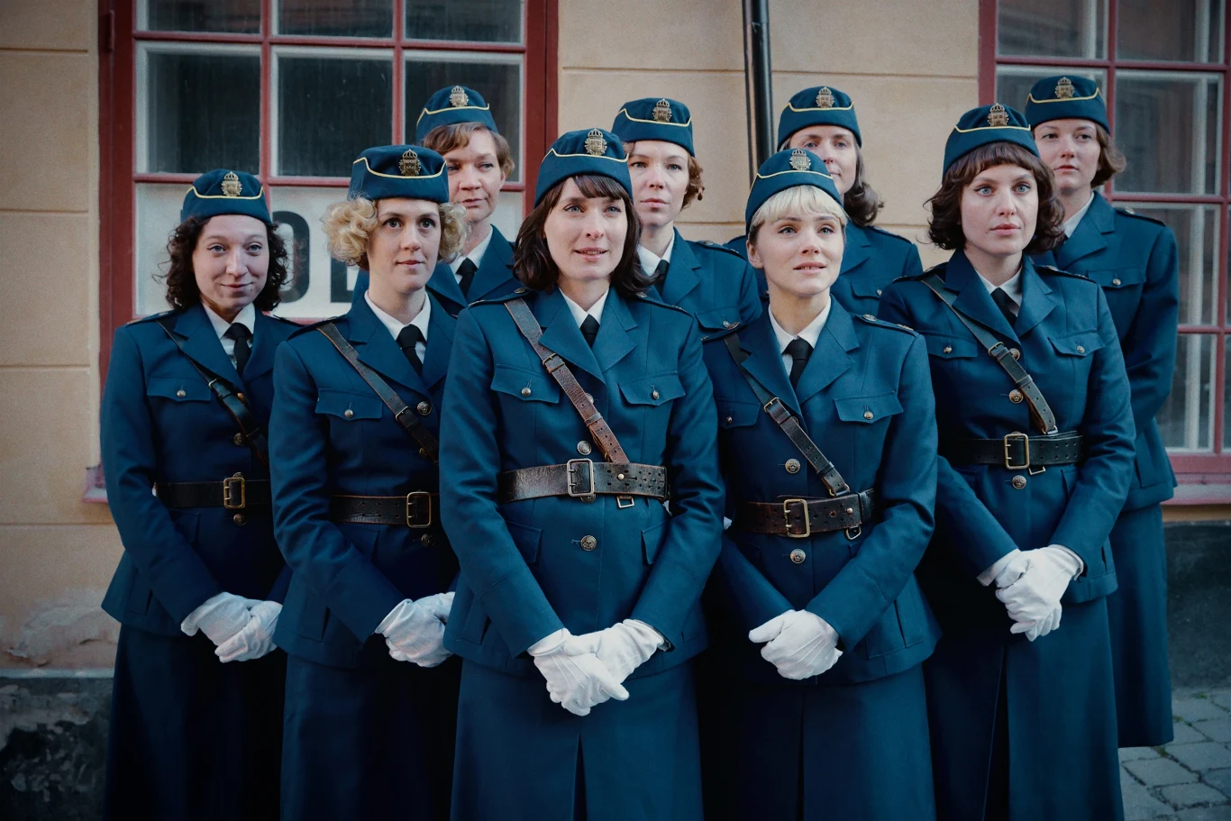 "Zmiana" Grupa kobiet w stylizowanych niebieskich mundurach służbowych, ustawionych w dwóch rzędach przed budynkiem z dużymi oknami, każda w białych rękawiczkach i wojskowych nakryciach głowy, tworzących oficjalną pozę.