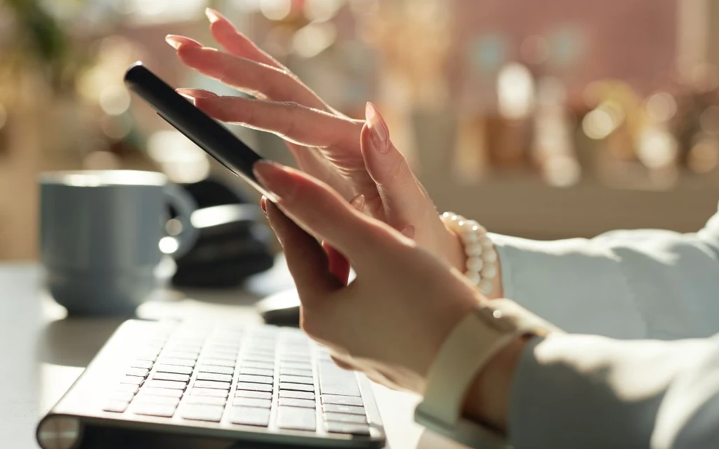 Zbliżenie na kobiece dłonie trzymające smartfona, przed klawiaturą komputerową; na dłoni widoczna bransoletka z pereł oraz zegarek, w tle kubek i rozmyte przedmioty biurowe.