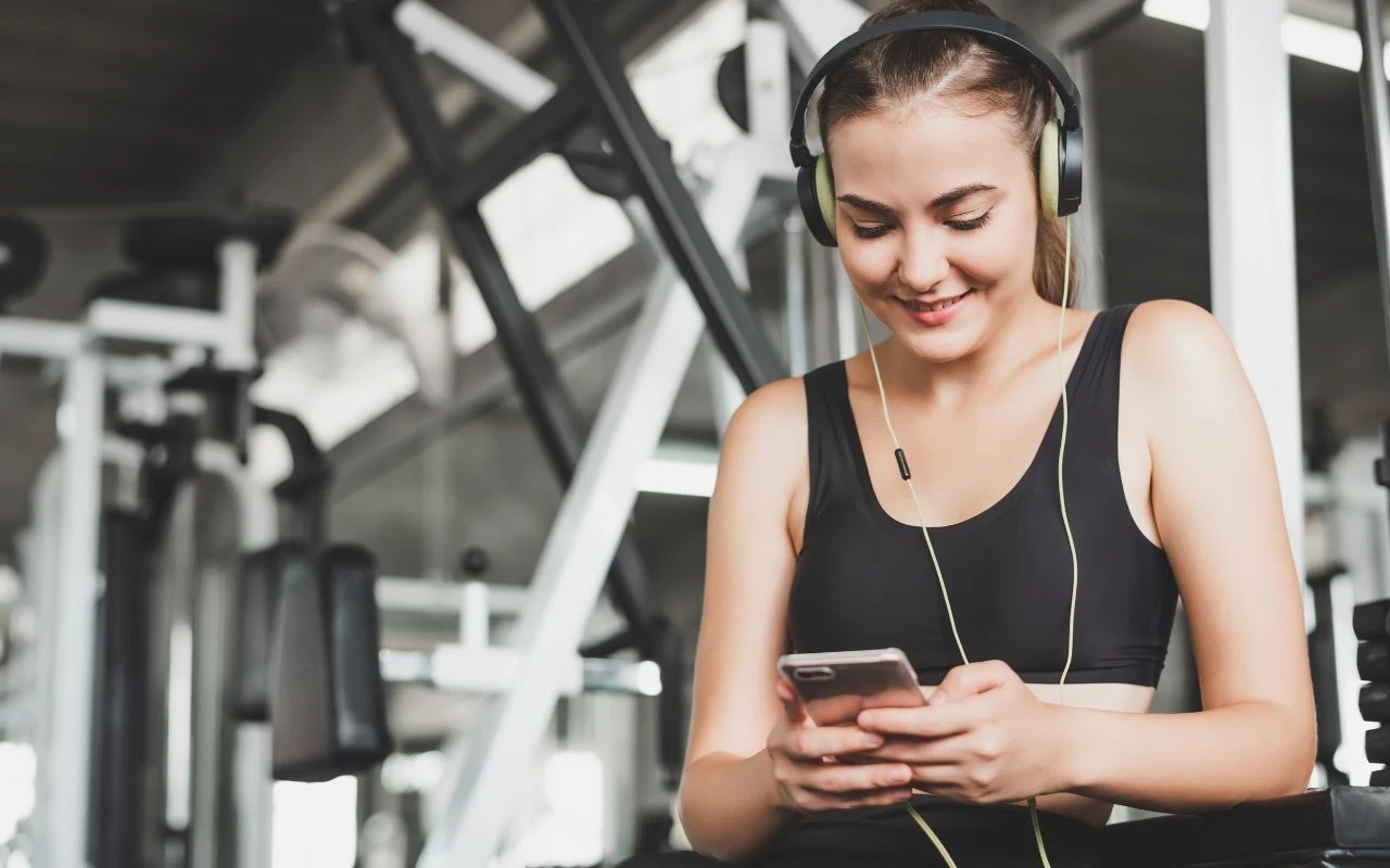 Muzyka na siłowni? Sprawdzone sposoby, aby uratować baterię Młoda kobieta w sportowym stroju siedzi na siłowni, słucha muzyki w słuchawkach i korzysta ze smartfona, otoczona sprzętem do ćwiczeń.