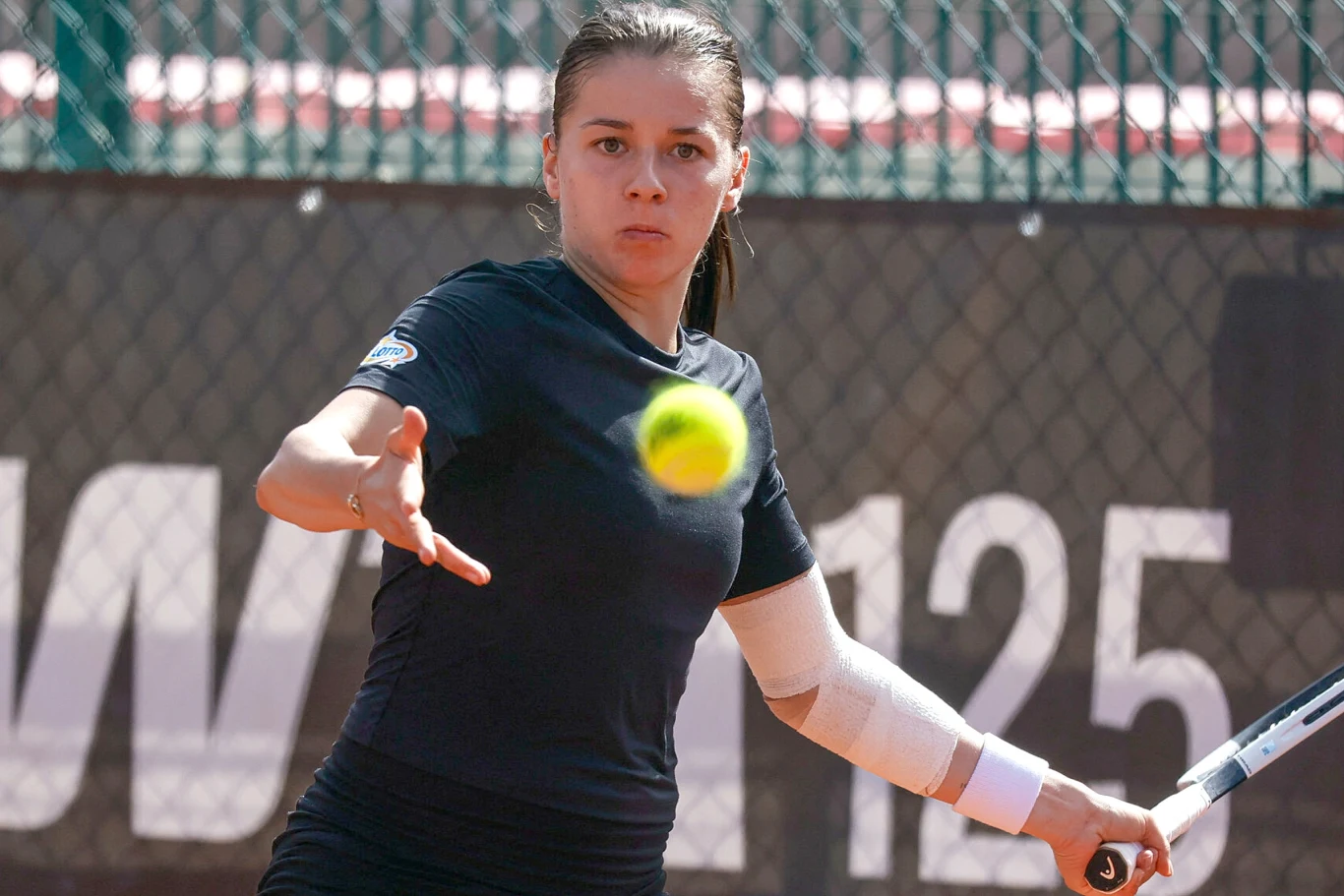 Tenisistka skupiona na odbiciu piłki podczas meczu na korcie, ujęcie dynamiczne z piłką unoszącą się w powietrzu, tło stanowi siatka i trybuny.