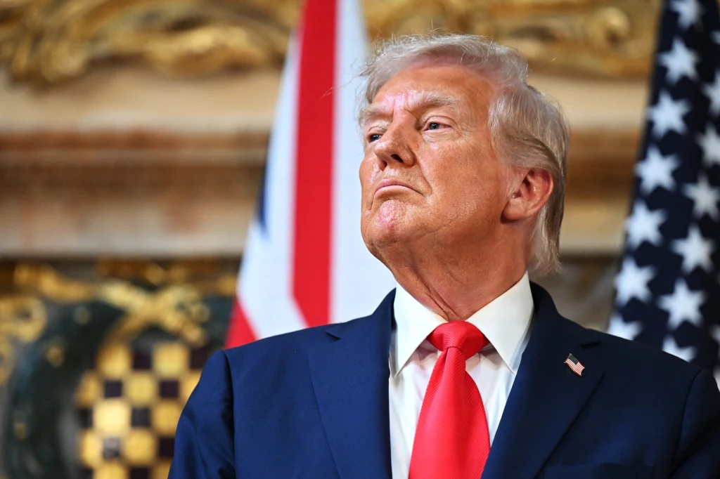 Prezydent Stanów Zjednoczonych Donald Trump Mężczyzna w garniturze i czerwonym krawacie stoi przed flagami USA i Wielkiej Brytanii w eleganckim wnętrzu, spoglądając poważnie w bok.