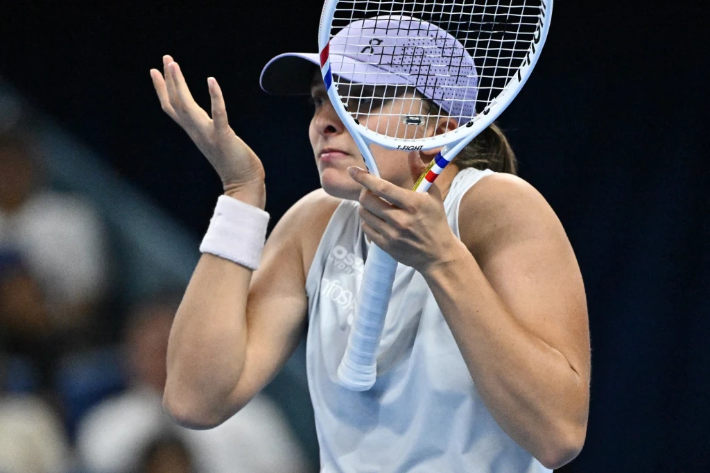 Tenisistka w sportowym stroju na korcie, unosi ramiona w geście niezrozumienia lub rozczarowania, trzymając rakietę blisko twarzy