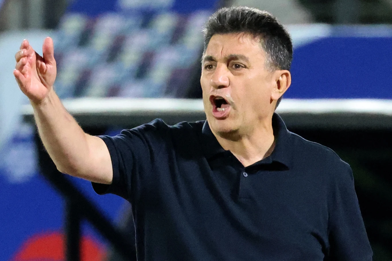 Amir Ghalenoei, selekcjoner piłkarskiej reprezentacji Iranu Mężczyzna w średnim wieku w ciemnej koszulce polo gestykuluje podniesioną ręką i ma otwarte usta, prawdopodobnie wydając polecenia lub komentując sytuację, w tle rozmyty stadion sportowy