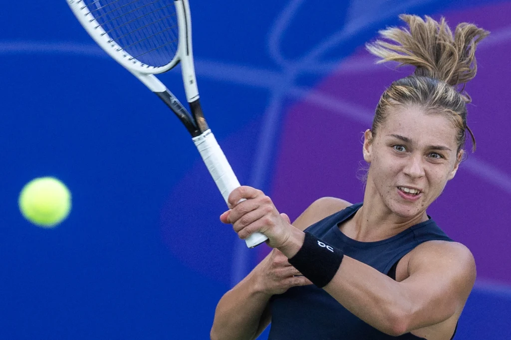 Maja Chwalińska Młoda kobieta w sportowym stroju odbija rakietą tenisową nadlatującą piłkę podczas meczu tenisowego na tle niebieskiego kortu.