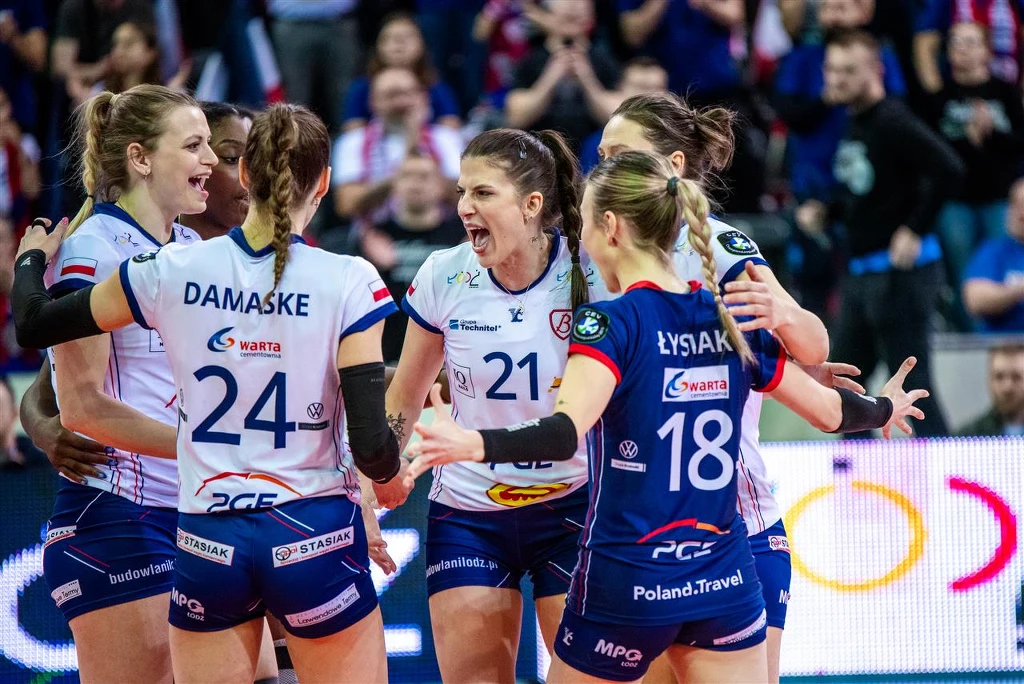 Grupa siatkarek w sportowych strojach świętuje zdobycie punktu na boisku, okazując entuzjazm i radość, wokół widoczna publiczność i atmosfera sportowego wydarzenia.