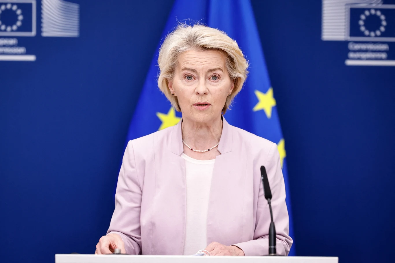 Ursulę von der Leyen czekają dwa wnioski o wotum nieufności Kobieta w oficjalnym stroju stoi za mównicą na tle flagi Unii Europejskiej oraz logotypów Komisji Europejskiej, przemawiając do mikrofonu.